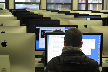 Room with many rows of tables. On each table there are several computer screens next to each others. A man sits in front of one of the computer screens and looks at it. The screen is turned on.